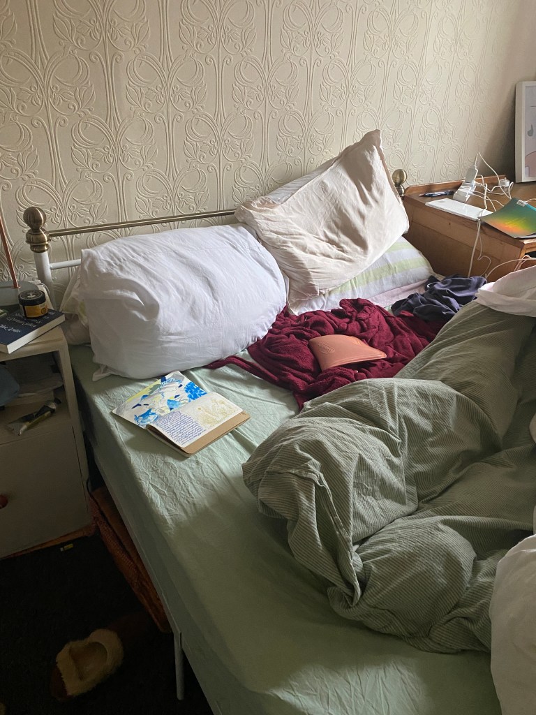 An unmade bed with mint green duvet showing an open notebook,hot water bottle and dressing gown